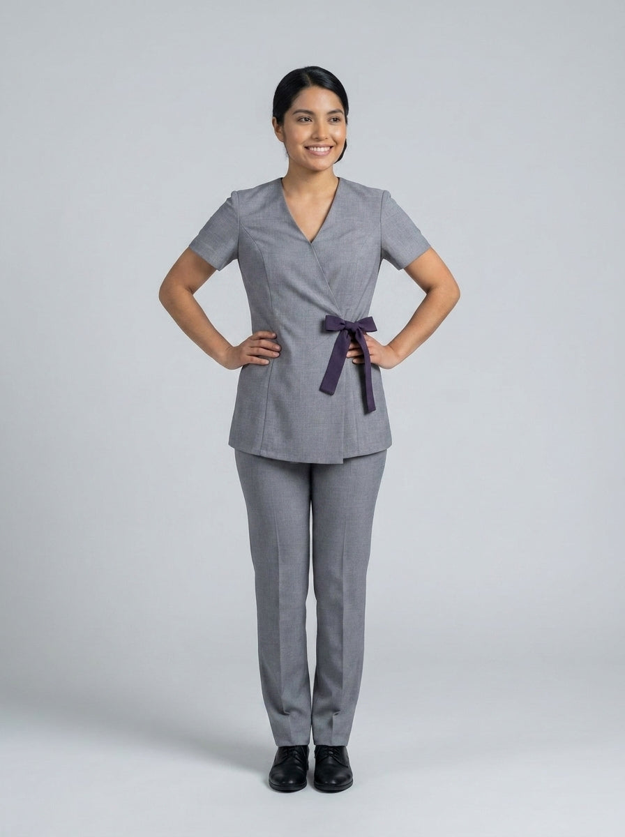 Dama con blusa gris cruzada de escote en V y lazo morado con pantalón gris para camarista de hotel.